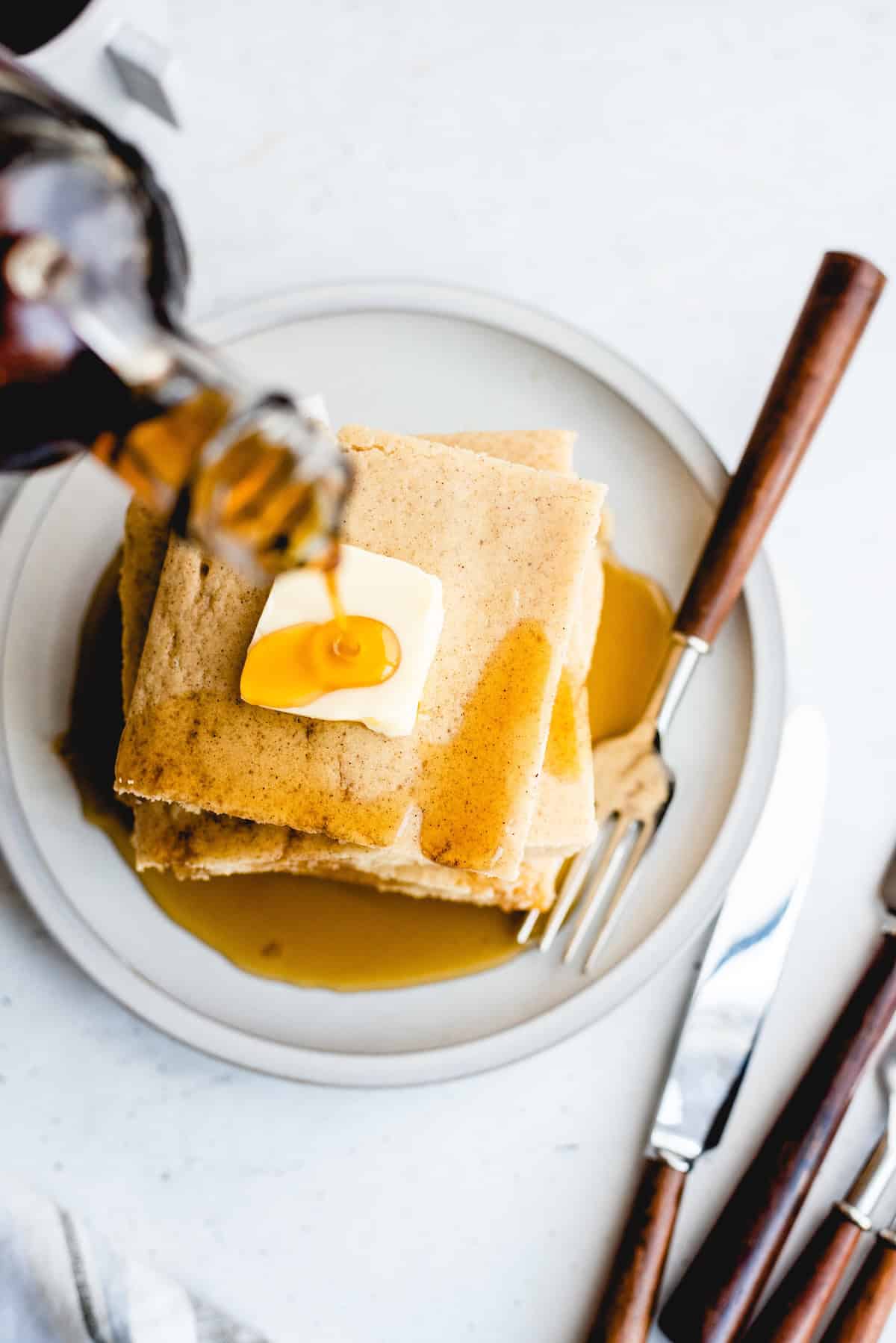 A stack of pancakes topped with a pat of butter and syrup being poured, served on a white plate with a fork and knife.