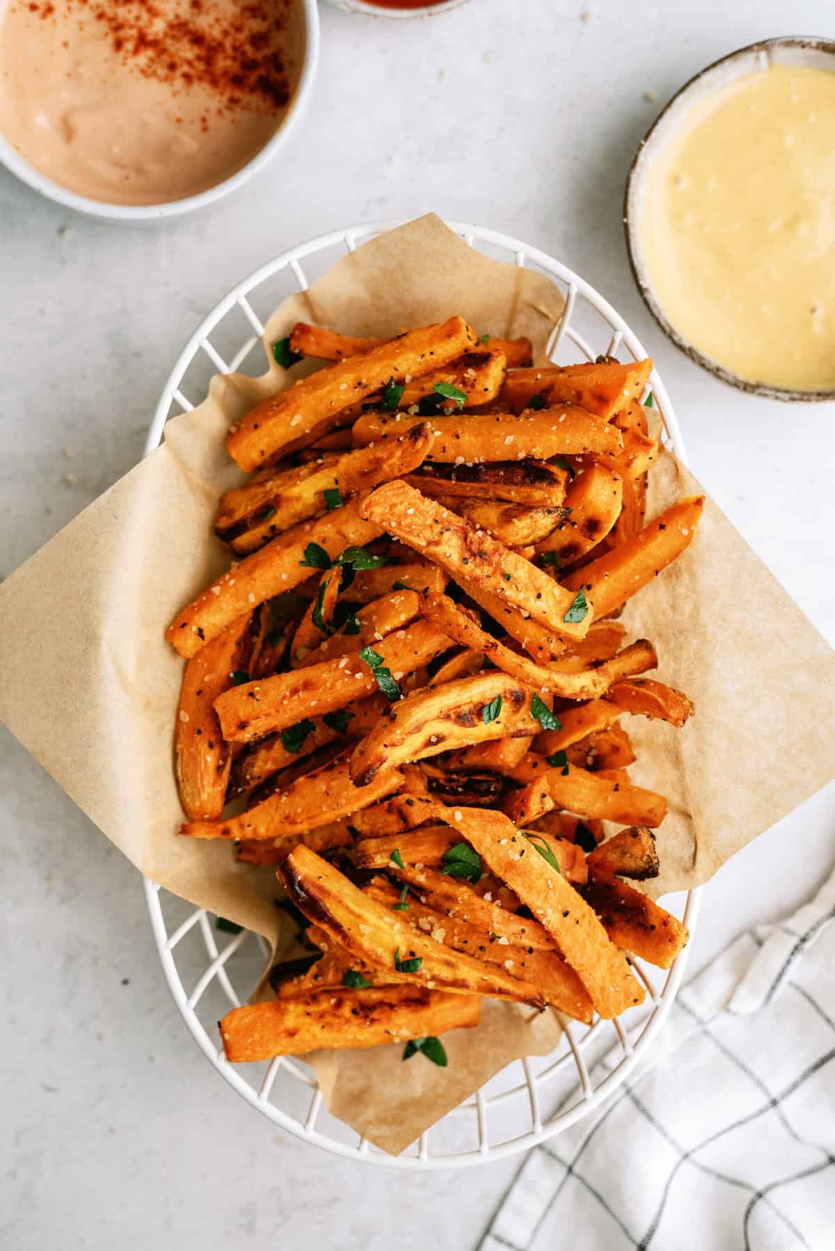 A basket of seasoned sweet potato fries garnished with herbs, placed on parchment paper, with two dipping sauces nearby.