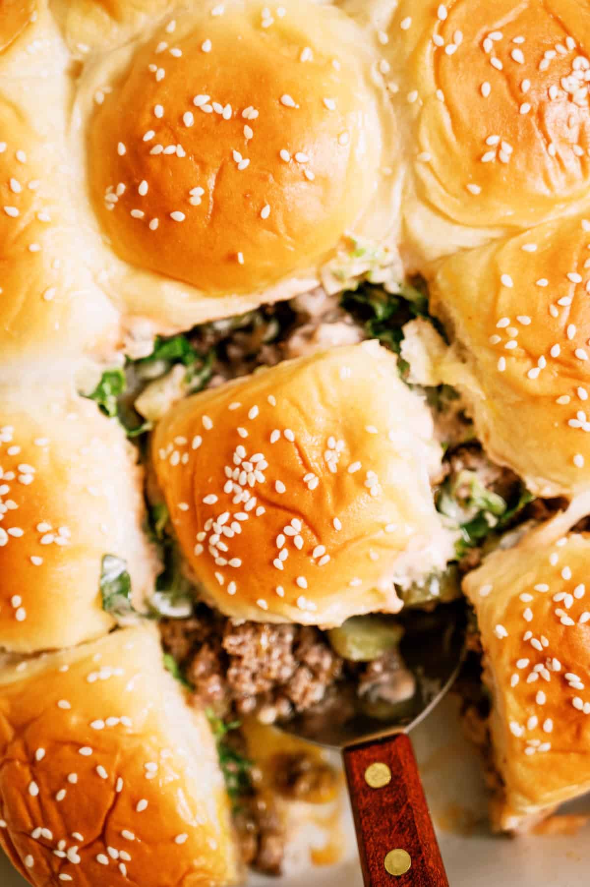 Close-up of a pan of pull-apart sliders with sesame seed buns, ground beef, lettuce, and melted cheese, with one slider being lifted out using a spatula.