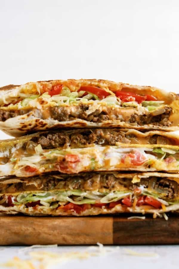 Two stacked halves of a quesadilla-style wrap filled with ground beef, shredded lettuce, diced tomatoes, cheese, and sauce, displayed on a wooden surface.