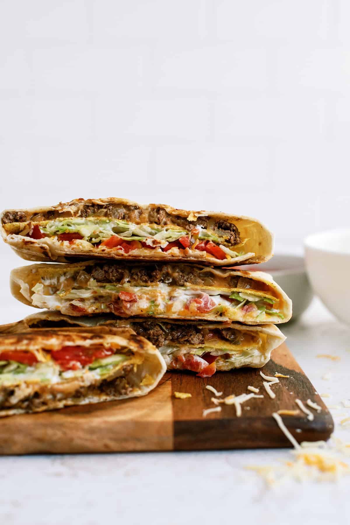 Three halved stuffed tortillas filled with ground meat, lettuce, tomato, cheese, and sauce are stacked on a wooden board with some shredded cheese scattered nearby.