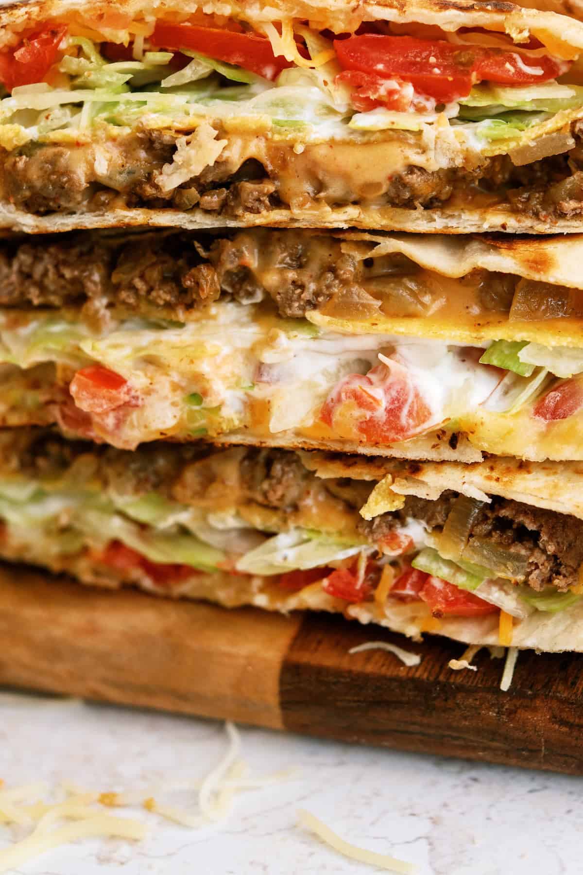 Close-up of stacked quesadillas filled with ground beef, melted cheese, lettuce, tomatoes, and tortillas on a wooden board.