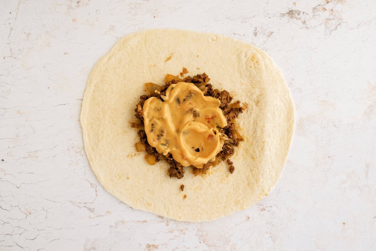 A flour tortilla topped with ground beef and a generous layer of melted cheese sauce sits on a white surface.