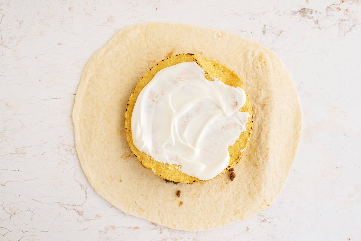 A piece of flatbread topped with a round fried patty and a layer of white sauce, placed on a light, textured surface.