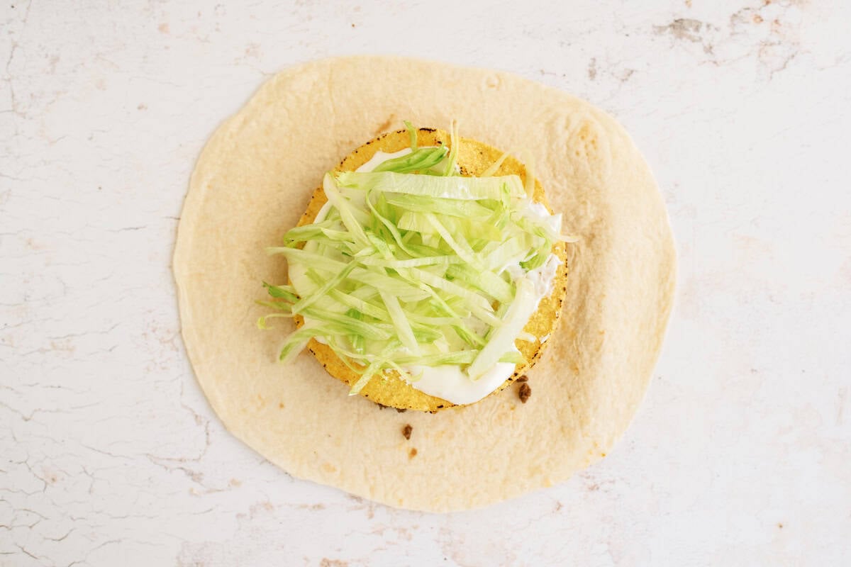 A flour tortilla topped with a round, breaded patty, shredded lettuce, and a white sauce on a light, textured surface.