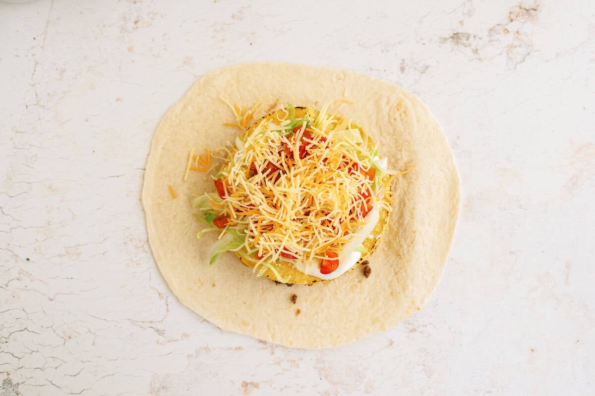 A flour tortilla topped with shredded lettuce, chopped tomatoes, shredded cheese, and a dollop of sour cream on a light, textured surface.