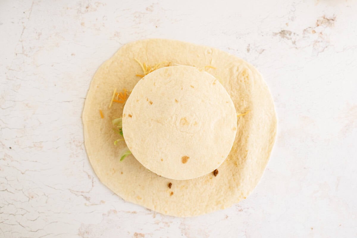 A small tortilla with fillings is placed on top of a larger tortilla on a light, textured surface.