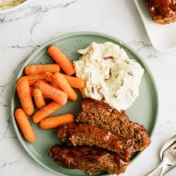 A green plate with glazed meatloaf slices, mashed potatoes with skins, and cooked baby carrots on a marble surface.