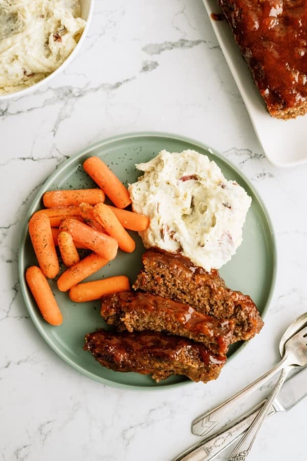 A green plate with glazed meatloaf slices, mashed potatoes with skins, and cooked baby carrots on a marble surface.