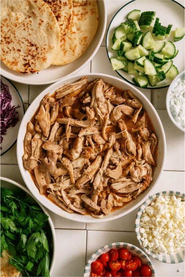 A bowl of Slow Cooker Chicken Shawarma surrounded by pita bread and toppings in bowls.