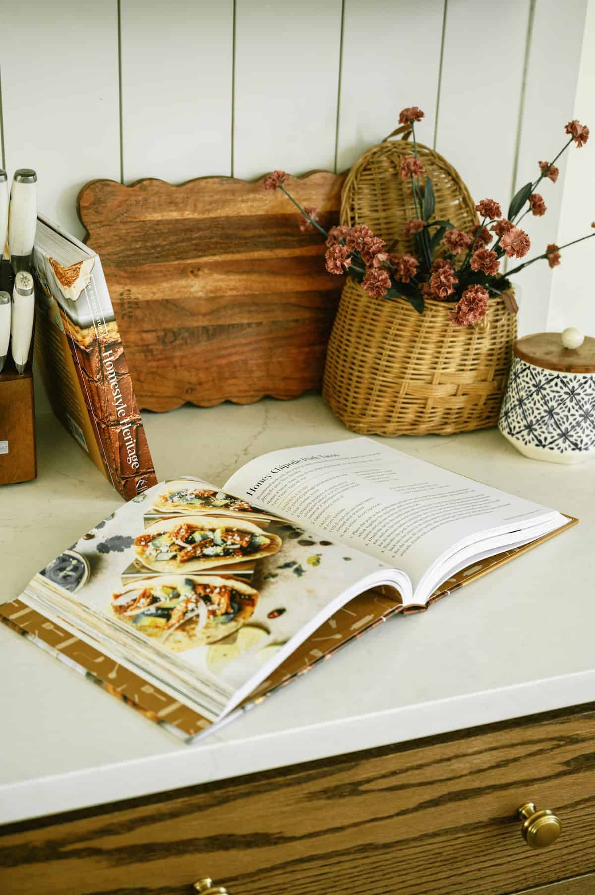 Open cookbook on kitchen counter with decorative baskets and flowers in the background.